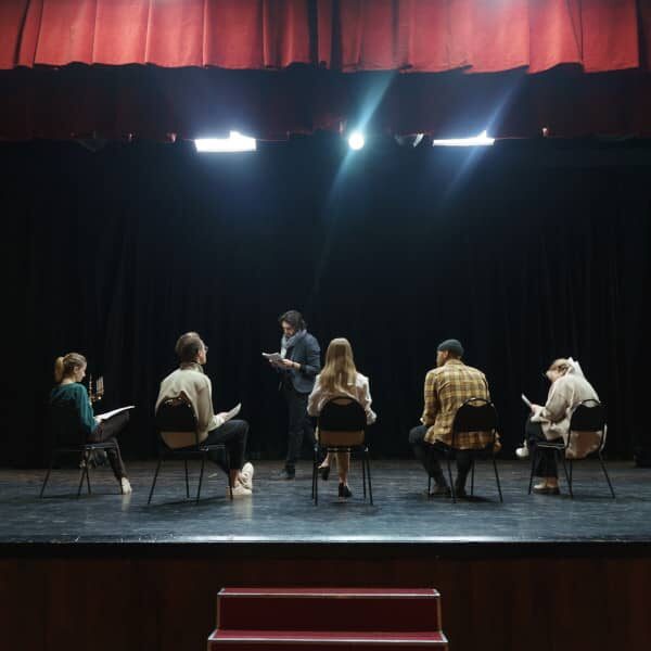 Fotografia di un gruppo di attori su un palcoscenico illuminato da 2 fari durante una sessione di prove con il regista.